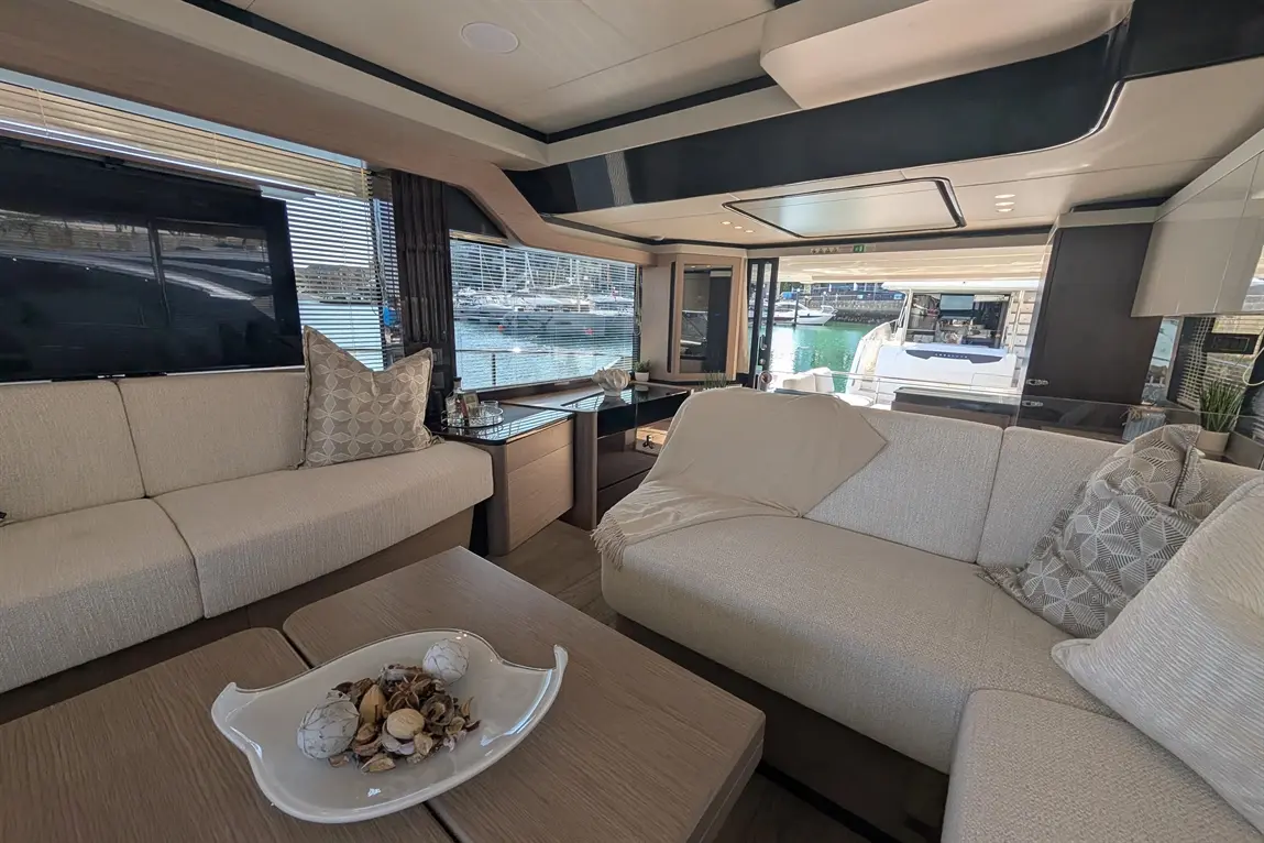 Luxurious yacht interior with beige sofas, a glass coffee table, and views of the marina through large windows.